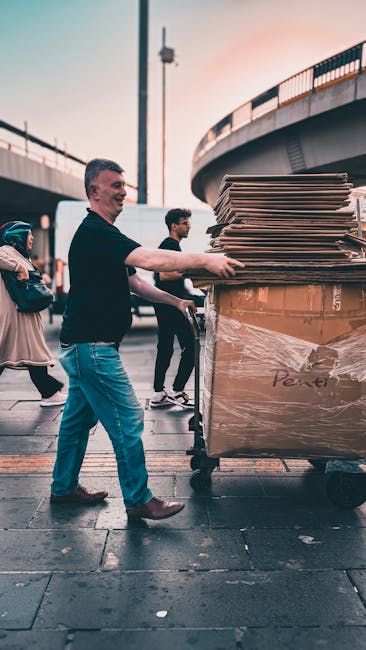 A man wearing a black shirt and blue jeans pushes a wheeled trolley loaded with flattened cardboard boxes and large sealed cardboard boxes wrapped in plastic on a moving dolly. He is outdoors, under a