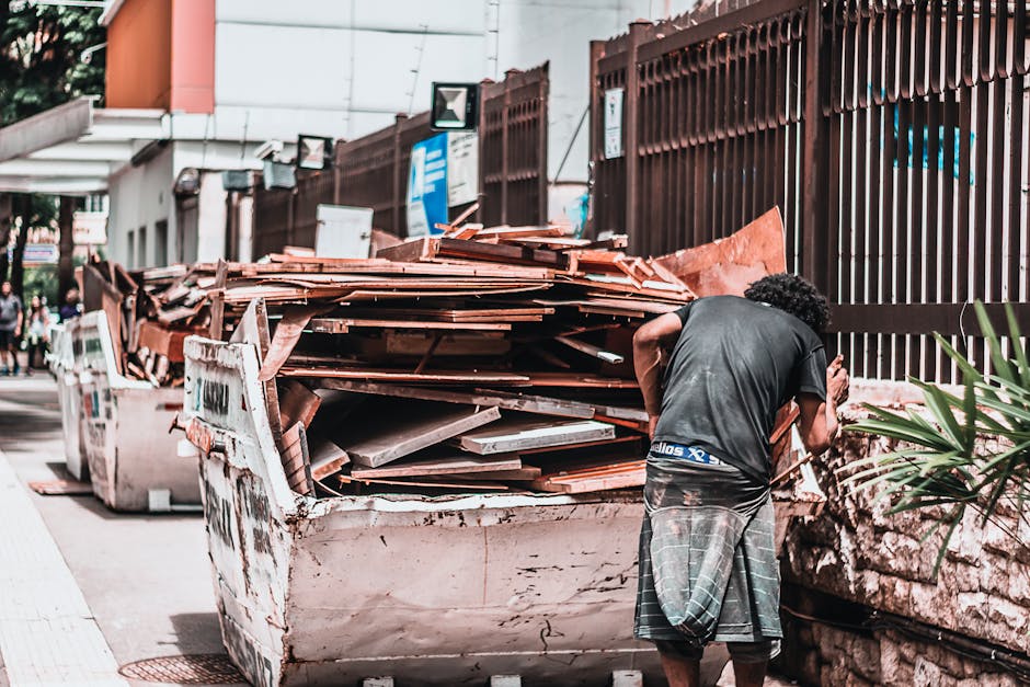 A person with curly black hair, wearing a dark t-shirt and grey checkered shorts, is standing next to a large, filled skip of construction debris and roofing tiles on a city street. The skip, position